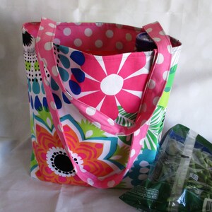 May include: A colorful tote bag with a floral pattern in pink, orange, blue, and green. The bag has pink polka dot lining and straps. A green bag of produce is visible next to the tote bag.
