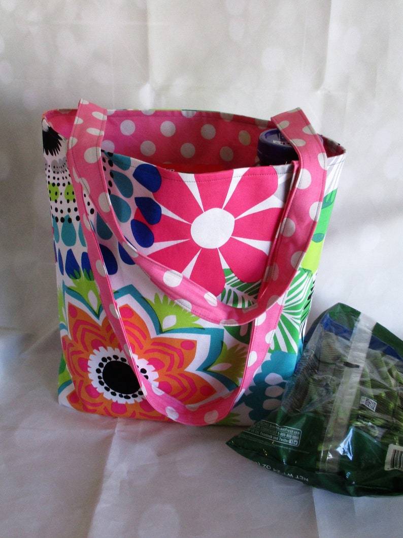 May include: A colorful tote bag with a floral pattern in pink, orange, blue, and green. The bag has pink polka dot lining and straps. A green bag of produce is visible next to the tote bag.