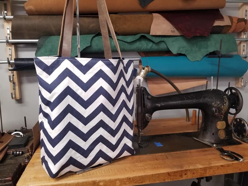 May include: A navy blue and white chevron pattern tote bag with brown leather handles. The bag is sitting on a wooden surface next to a vintage black sewing machine. Rolls of leather are visible in the background.