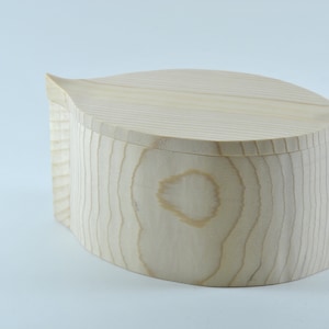 Puede incluir: Una caja de madera clara con una tapa redondeada en forma de hoja. La caja está hecha de madera natural con veta visible.