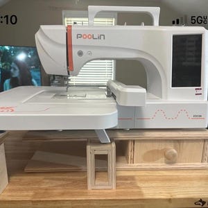 May include: A white Poolin sewing machine with an orange accent, resting on a wooden table. The machine has a digital display and is mounted on a wooden stand with a drawer. The table is made of wood and has the word "Husky" printed on it.