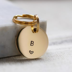May include: A gold-coloured keyring with a circular pendant. The pendant displays the letter "B" and a heart symbol. The keyring has a double ring for attaching keys.