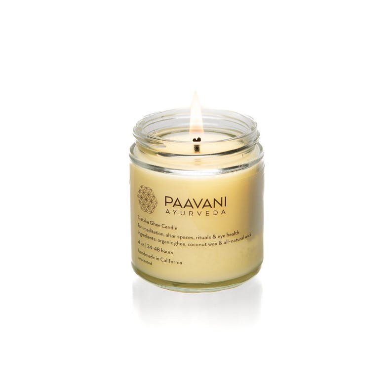 May include: A glass jar candle with a golden yellow wax and a single wick. The jar has a label that reads "PAAVANI AYURVEDA" and "Troika Ghee Candle". The label also includes information about the candle's use for meditation, altar spaces, rituals, and eye health. The candle is made with organic ghee, coconut wax, and all-natural ingredients. It burns for 24-48 hours and is handmade in California.