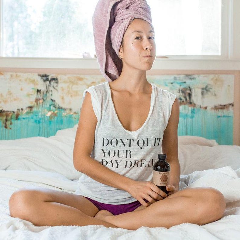 Op de afbeelding: Een vrouw in een wit tanktop met de tekst "Don't Quit Your Day Dream" zit op een bed met een flesje vloeistof in haar hand. Ze heeft een roze handdoek om haar hoofd gewikkeld.