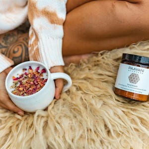 May include: A white mug filled with a pink and white herbal tea blend, with dried rose petals floating on top. A brown jar with a black lid and white label reads "Paavani Ayurveda Shatavari" on a fluffy white rug.
