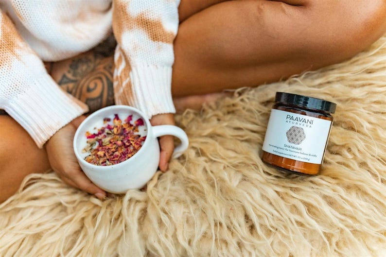 May include: A white mug filled with a pink and white herbal tea blend, with dried rose petals floating on top. A brown jar with a black lid and white label reads "Paavani Ayurveda Shatavari" on a fluffy white rug.