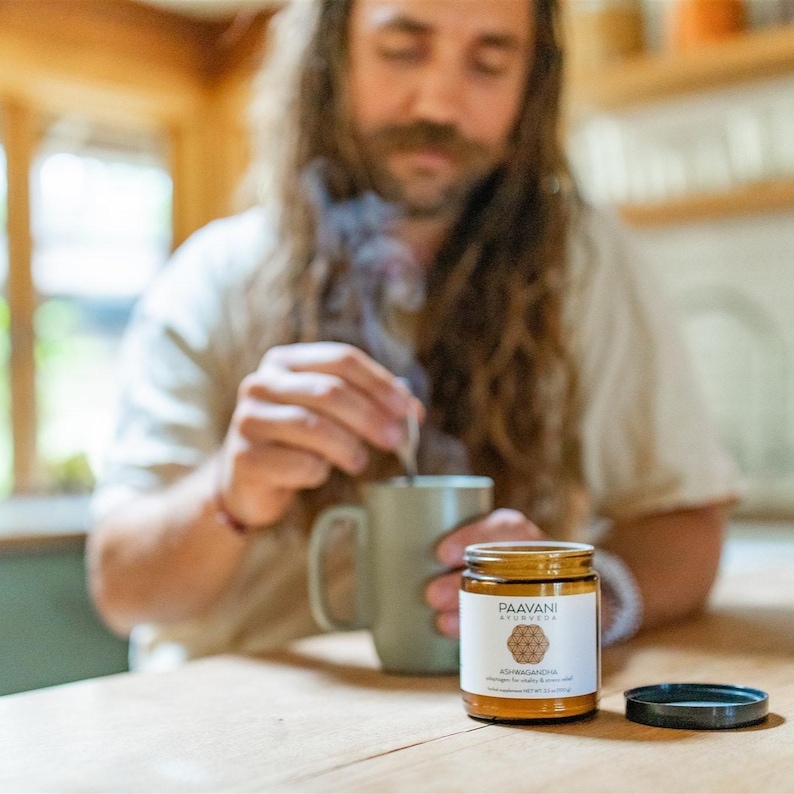 Puede incluir: Un tarro de vidrio marr&oacute;n con tapa dorada y etiqueta que dice "Paavani Ayurveda Ashwagandha Chai" con un dise&ntilde;o geom&eacute;trico. El tarro est&aacute; sobre una mesa de madera delante de una persona que sostiene una taza.
