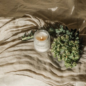 May include: A white candle in a glass jar with a black label that reads "PAAVANI AYURVEDA". The candle is lit and the flame is visible. The candle is sitting on a beige fabric surface with green leaves and flowers.