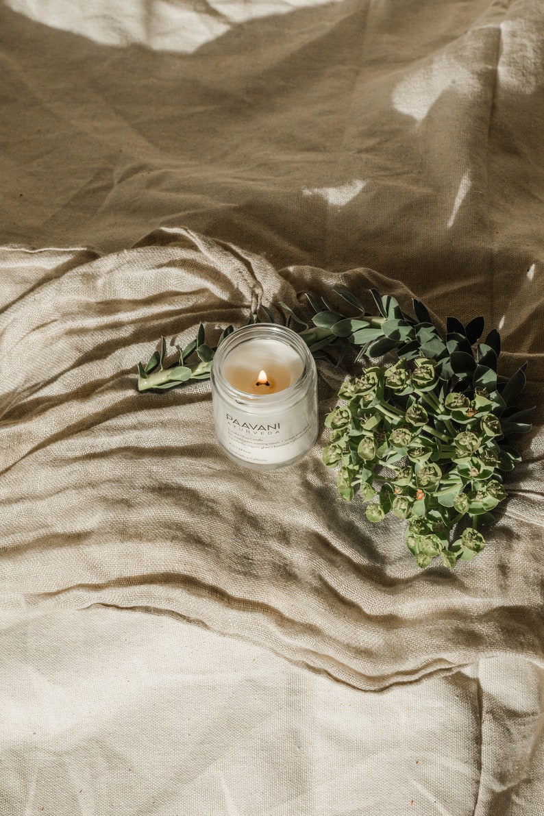 May include: A white candle in a glass jar with a black label that reads "PAAVANI AYURVEDA". The candle is lit and the flame is visible. The candle is sitting on a beige fabric surface with green leaves and flowers.