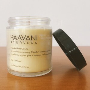 May include: A clear glass jar with a yellow candle inside. The jar has a black label with the text "PAAVANI AYURVEDA" and the words "Trataka Ghee Candle" and "4 oz. | 24 hour" printed on it. The candle is handmade in California.