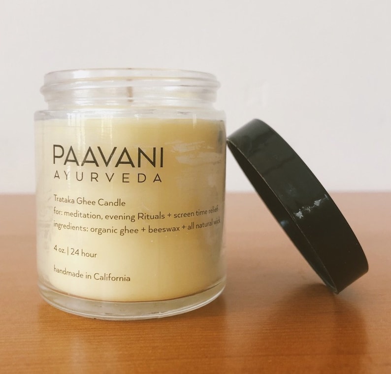 May include: A clear glass jar with a yellow candle inside. The jar has a black label with the text "PAAVANI AYURVEDA" and the words "Trataka Ghee Candle" and "4 oz. | 24 hour" printed on it. The candle is handmade in California.