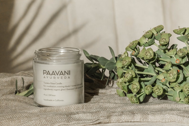 May include: A white candle in a clear glass jar with a white label that reads "Paavani Ayurveda" and "Trataka Ghee Candle" for meditation, evening rituals + screen time with all natural ingredients: organic ghee + beeswax + all-natural essential oils. 4 oz | 24 hour handmade in California.
