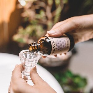 Op de afbeelding: Een bruine glazen fles met een etiket met de tekst "PAAVANI" en een geometrisch ontwerp. De fles wordt in een heldere glazen beker gegoten.