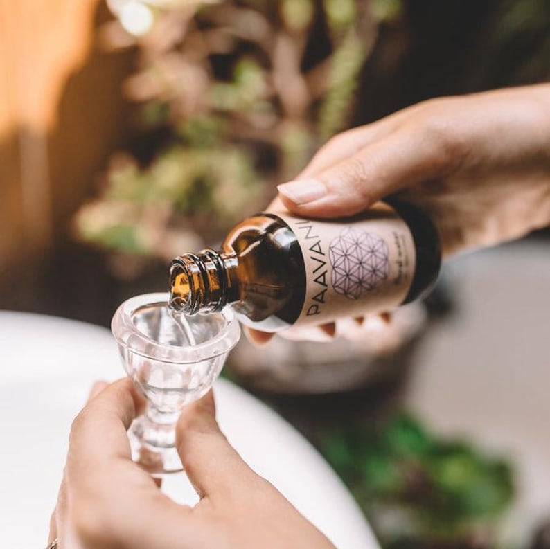 Op de afbeelding: Een bruine glazen fles met een etiket met de tekst "PAAVANI" en een geometrisch ontwerp. De fles wordt in een heldere glazen beker gegoten.