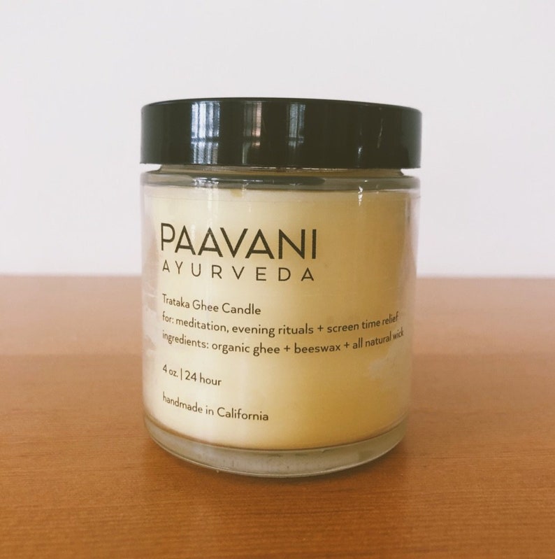 May include: A glass jar candle with a black lid and a yellow label that reads "PAAVANI AYURVEDA Trataka Ghee Candle for: meditation, evening rituals + screen time relief ingredients: organic ghee + beeswax + all natural wick 4 oz. / 24 hour handmade in California"