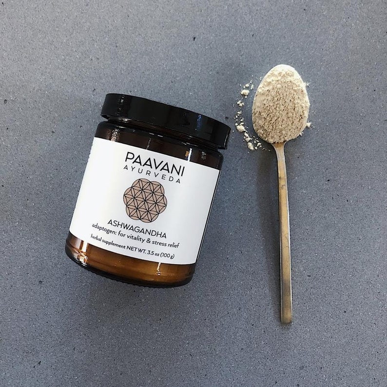 Puede incluir: Un tarro de vidrio marr&oacute;n con tapa negra que contiene un polvo marr&oacute;n claro. El tarro tiene una etiqueta blanca con el texto "PAAVANI AYURVEDA ASHWAGANDHA adaptogen: for vitality & stress relief herbal supplement NET WT. 3.5 oz (100 g)". Una cuchara de oro con una cucharada llena de polvo est&aacute; al lado del tarro.