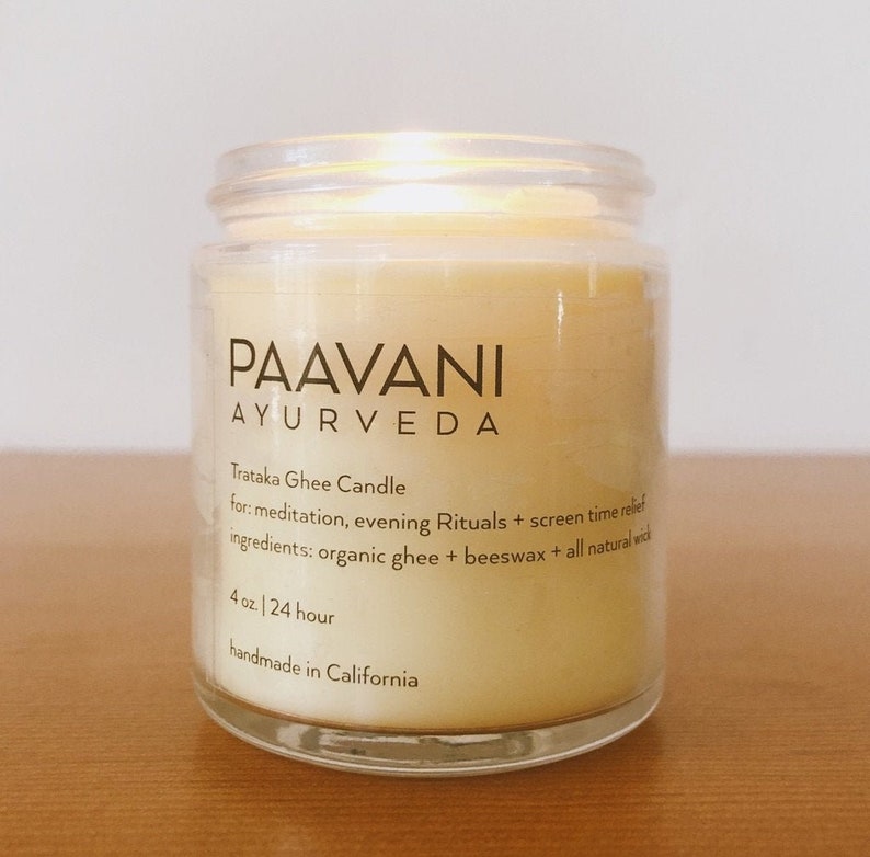 May include: A clear glass jar with a yellow candle inside. The label reads "PAAVANI AYURVEDA" and "Trataka Ghee Candle" with details about the candle's purpose and ingredients. The candle is 4 oz and burns for 24 hours. It is handmade in California.