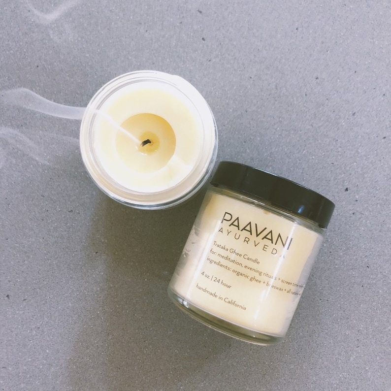 May include: A glass jar with a black lid containing a white candle. The jar has a label that reads "PAAVANI AYURVEDA Trataka Ghee Candle for meditation, evening ritual + screen time. Ingredients: organic ghee + beeswax + all natural essential oils. 4 oz. | 24 hour handmade in California."