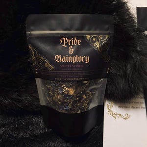 Puede incluir: Una bolsa negra vertical que contiene té a granel con una ventana transparente. La etiqueta presenta un texto dorado ornamentado que dice Pride & Vainglory, Night Excision, Hand Blended Teas, Made in Portland OR y la URL www.etsy.com/shop/nightexcision.