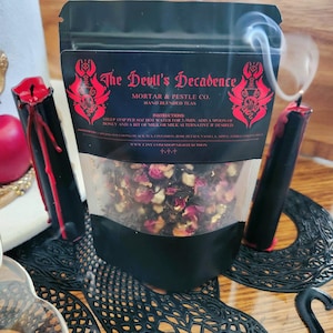 May include: A black pouch of "The Devil's Decadence" hand-blended teas, with visible dried rose petals and other ingredients. The pouch is flanked by two black candles with red wax and a decorative black coaster. Smoke rises from the scene.