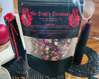 Pode incluir: Uma bolsa preta de chás misturados à mão "The Devil's Decadence", com pétalas de rosa secas visíveis e outros ingredientes. A bolsa é ladeada por duas velas pretas com cera vermelha e um porta-copos preto decorativo. A fumaça sobe da cena.