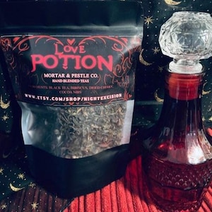 May include: A black pouch of "Love Potion" tea from Mortar & Pestle Co. sits next to a red glass bottle with a crystal stopper. The tea bag lists ingredients: black tea, hibiscus, dried cherry, and cocoa nibs.