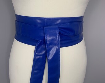 Pode incluir: Um cinto largo de couro sintético azul com uma faixa longa que pode ser amarrada em um laço. O cinto é mostrado amarrado em torno de um manequim branco.