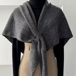 May include: A grey wool cape scarf with a knotted front. The scarf is draped over a black turtleneck and a beige skirt. The scarf's texture appears soft and warm, suitable for cooler weather. The overall style is elegant and minimalist.