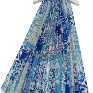 Puede incluir: Bufanda estampada azul y blanca con diseño floral. La bufanda está colgada de una percha blanca. La bufanda tiene una variedad de tonos azules, incluyendo azul claro y oscuro, con un fondo blanco. La bufanda es de un material ligero.