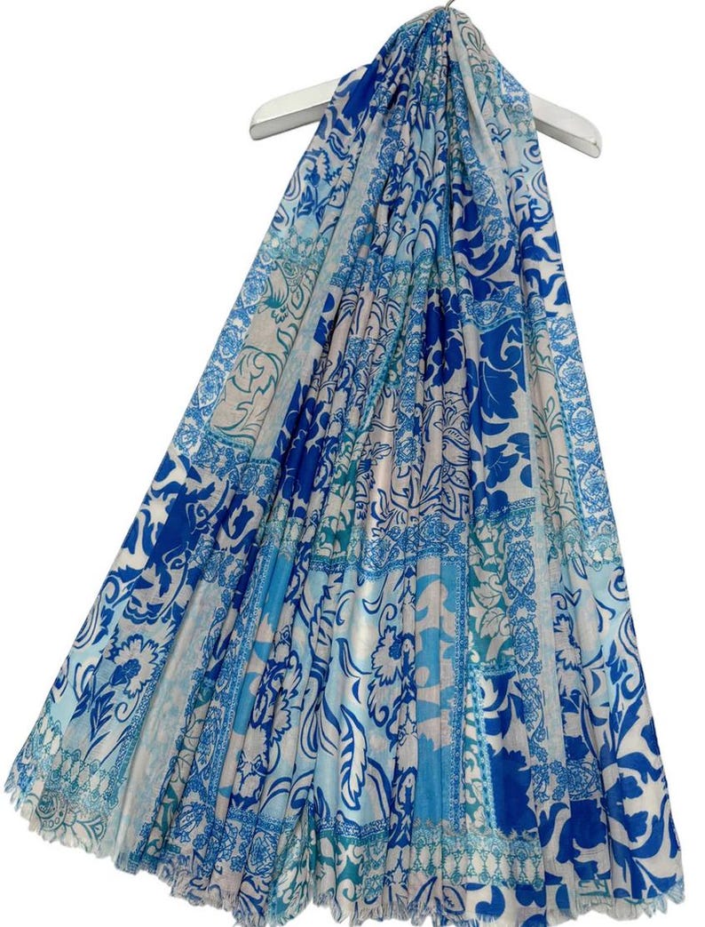 May include: A blue and white patterned scarf with a floral design. The scarf is draped over a white hanger. The scarf has a variety of blue shades, including light and dark blue, with a white background. The scarf is made of a lightweight material.