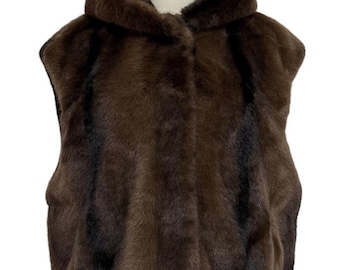 May include: A brown faux fur vest with a hood. The vest is displayed on a mannequin and has a soft, textured appearance. The hood is lined with a darker shade of brown, and the vest has a button closure.