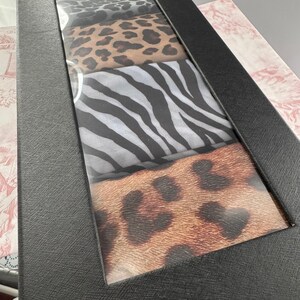 May include: A black rectangular box with a clear window showing four patterned fabrics. The patterns include leopard print, zebra stripes, and other animal prints. The box is on a patterned surface.
