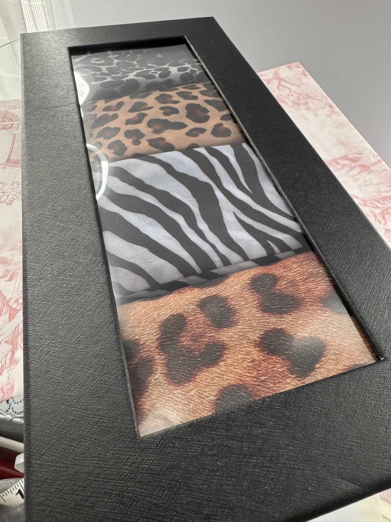 May include: A black rectangular box with a clear window showing four patterned fabrics. The patterns include leopard print, zebra stripes, and other animal prints. The box is on a patterned surface.