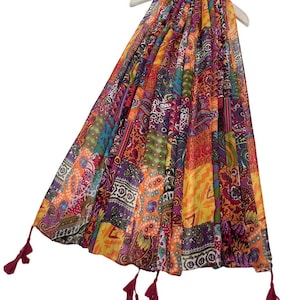 Può includere: Una sciarpa patchwork colorata con una varietà di motivi e colori, tra cui arancione, viola, giallo e turchese. La sciarpa ha un tessuto leggero e fluido ed è adornata con nappe bordeaux alle estremità. È esposta su una gruccia bianca.