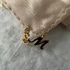 May include: A gold-coloured charm featuring the letter "M" in black enamel, attached to a beige fabric with a stitched edge. The charm is set against a light grey, textured background.