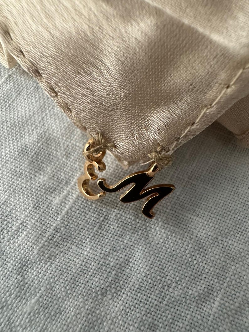 May include: A gold-coloured charm featuring the letter "M" in black enamel, attached to a beige fabric with a stitched edge. The charm is set against a light grey, textured background.