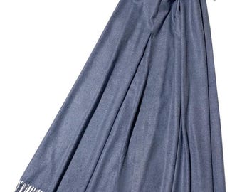 May include: A denim blue scarf with fringe detail. The scarf is draped over a white hanger, showcasing its length and texture. The fabric appears soft and lightweight, suitable for cooler weather.