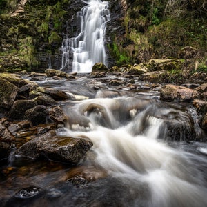 May include: A long exposure shot of a waterfall flowing over dark rocks. The water appears blurred, creating a sense of movement. Green moss and vegetation surround the waterfall. A logo is in the top left corner.