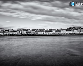 May include: A black and white photograph of a row of houses along a waterfront with a cloudy sky above.