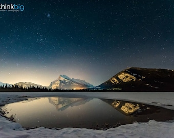 May include: A night sky filled with stars over a frozen lake with a mountain range in the background. The reflection of the mountains and stars can be seen in the lake.