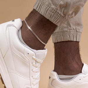 Può includere: Una cavigliera a catena d'argento indossata sopra una sneaker bianca. La cavigliera è un delicato design a catena. La persona indossa pantaloni beige. Lo sfondo è di colore beige.
