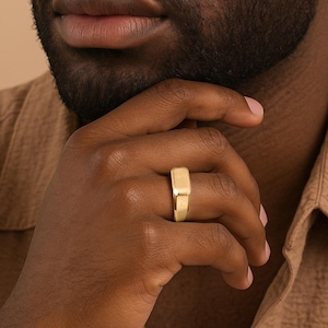 Puede incluir: Un anillo de oro con una parte superior rectangular se lleva en un dedo. El anillo tiene un diseño simple y moderno y un acabado pulido. El anillo es una joya.