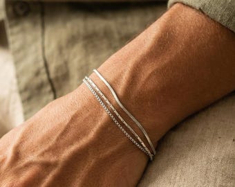 Pode incluir: Um conjunto de duas pulseiras de tom prateado no pulso. Uma pulseira apresenta um design de corrente de cobra plana e flexível, enquanto a outra é uma corrente de caixa fina e delicada. Ambas as peças têm um acabamento metálico polido.