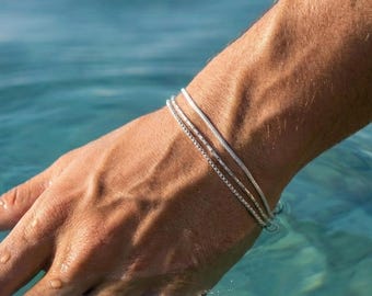 Pode incluir: Um conjunto de duas pulseiras finas de metal prateado usadas no pulso. As pulseiras apresentam um design delicado de elos refletivos que captam a luz, criando um brilho sutil na pele enquanto repousam logo acima da superfície da água.