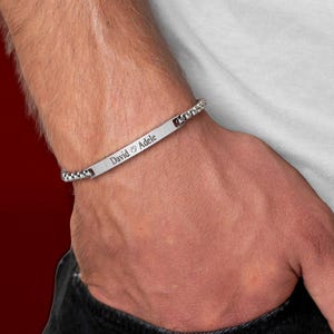 May include: A silver bracelet engraved with "David & Adele". The bracelet has a rectangular plate with the names and a chain design on each side. The bracelet is worn on a wrist.