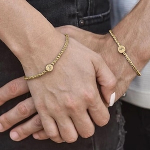 May include: Gold-colored braided chain bracelets. Each bracelet features a circular charm with a design. The bracelets are worn on wrists. The background is blurred, with a person wearing a black denim jacket.