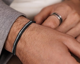 May include: A black and silver beaded bracelet with a thin white stripe is worn on a wrist. A silver ring with a black stripe is on the adjacent finger. The bracelet is a stylish accessory.
