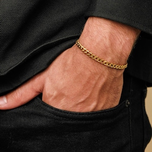 Pode incluir: Uma pulseira de corrente de elos dourada. A pulseira é usada no pulso, com a mão no bolso. A pulseira é feita de pequenas elos interligados. O fundo é uma jaqueta preta e calças pretas.