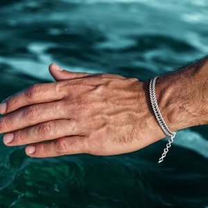 Op de afbeelding: Een zilveren kettingarmband met een gevlochten ontwerp. De armband wordt om de pols gedragen.