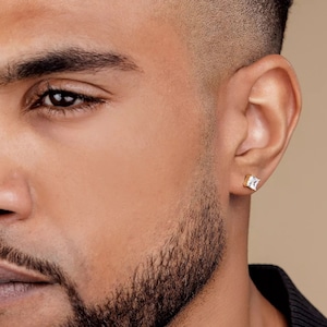 May include: A close-up of a man's ear with a gold square-shaped earring with a clear stone in the center.